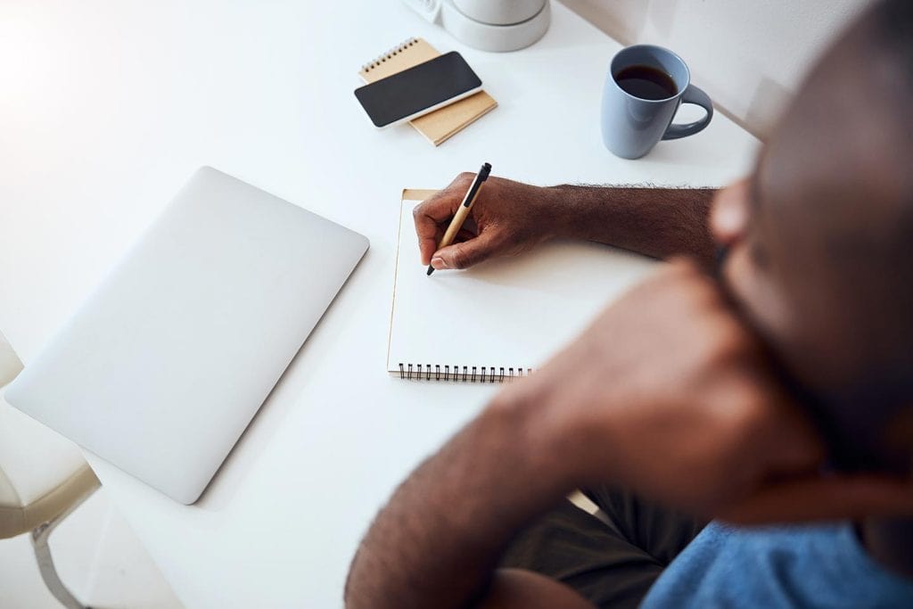 Man sits at desk with closed laptop, writing in a notebook.